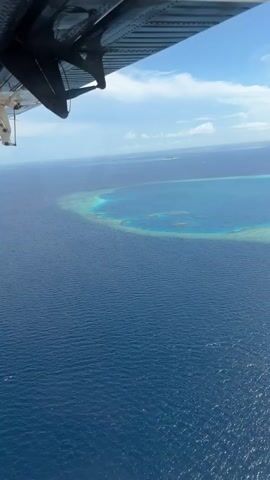 An aerial view of a vast expanse of deep blue ocean with a bright turquoise coral reef visible near the center. The reef appears as a shallow, circular formation with lighter blue patches within it, indicating submerged structures. In the distance, a small, uninhabited island with lush vegetation is visible. The sky is a clear blue with scattered white clouds. The bottom left corner of the frame shows a portion of an aircraft wing or fuselage, suggesting the footage was captured from a plane. The water exhibits a gentle ripple pattern, indicating calm conditions. No people or other objects are discernible in the water or on the island.