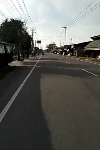 The video shows a first-person view moving down a black asphalt road with a white line marking the edge.  On either side of the road are various buildings, some appearing to be stores or small businesses, and others seemingly residential. Power lines are strung overhead, supported by utility poles.  A few vehicles, including motorcycles and a truck, are visible in the distance. The sky is overcast, casting long shadows across the road.