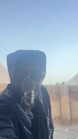 A person wearing a black hoodie and sunglasses with a gold chain is shown from the chest up. They are standing against a light brown wall with a clear sky in the background. The person moves their hands in a gesture.
