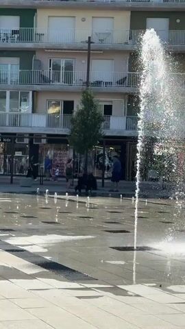 The video shows a street scene in front of buildings with balconies. In the foreground, there is a splash pad with water jets creating a spray. People and dogs are visible on the sidewalk. On the left, a woman and a dog are standing near the splash pad. A man is standing next to them. Behind them, there are shop windows and a tree. Further to the right, a white car is parked. The water from the splash pad is reflecting the light and creating a shimmering effect on the ground. The overall impression is a sunny day in an urban environment.
