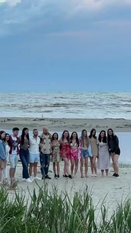 A wide shot captures a group of around a dozen people standing on a sandy beach. They are facing the camera, posing for a photo. The group consists of men and women of varying ages, dressed in casual summer attire. Behind them, a body of water stretches out to the horizon under a dramatic sky filled with swirling clouds, tinged with the colors of sunset. Tall green grass and reeds grow in the foreground, partially obscuring the lower part of the frame. As the camera zooms out, a person wearing a green shirt and black pants is seen taking a picture of the group with their phone. There is another person walking away on the beach in the distance. The atmosphere is casual and celebratory.
