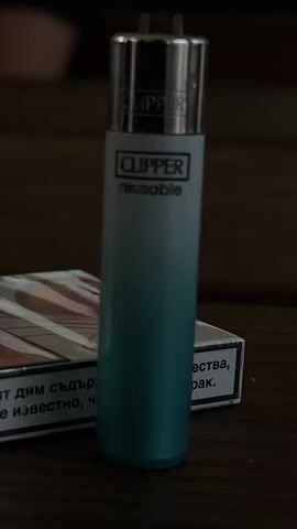 The image presents a close-up, static view of a Clipper lighter and a partially visible pack of cigarettes, resting on a dark wooden surface.

The lighter, centrally positioned and standing upright, features a metallic silver top and a cylindrical body with an ombré color transition. It is white at the top, gradually fading into a teal or turquoise shade towards the base. The brand name "CLIPPER" is visible both on the metallic top and prominently on the white section of the lighter's body, where it is followed by the word "reusable" in smaller print.

Behind the lighter, a pack of cigarettes is partially visible and slightly out of focus. Its design shows a white and orange pattern, but a significant portion of its lower half is covered by a white health warning label. This label contains fragmented text in Cyrillic script. Partially legible phrases include "ДИМ СЪДЪРЖА" (smoke contains) and "РАК" (cancer), which are common elements of tobacco health warnings.

Both items are situated on what appears to be a dark, polished wooden table or surface. The background behind the objects is dark and heavily blurred, making any further details indistinguishable. The overall lighting is dim, suggesting an indoor setting with subdued illumination.

No people are visible, and no activity is taking place; the scene is a still life of the objects. The Cyrillic text on the cigarette pack indicates a location in a Slavic-speaking country, consistent with Bulgaria.