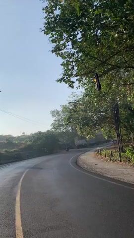 A camera mounted on a moving vehicle captures a view of a paved road winding through a natural landscape. The road has a yellow line marking its center and a white line on its right edge. On the left side of the road, there is a grassy embankment with trees and some small buildings. On the right side, there are more trees and a wooden structure. Several vehicles are visible on the road, including a truck and at least two motorcycles with riders. The sky is clear and blue, indicating daytime. The overall scene suggests a rural or semi-rural setting with active road traffic.