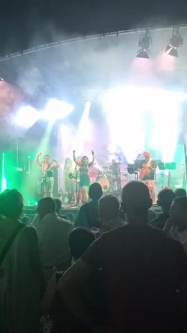 A band is performing on a stage illuminated by bright white and green lights. The band consists of at least four members: a female singer in the center, two guitarists on either side, and a drummer in the back. The singer is wearing a dark top and shorts, and her arms are raised in the air. The guitarists are holding their instruments and appear to be playing. The drummer is seated behind a drum kit. The stage is set up outdoors, with a dark sky visible above the stage structure. A crowd of people is watching the performance from the foreground, with their backs to the camera. Some members of the crowd are illuminated by the stage lights, while others are in shadow. The scene is filled with atmospheric smoke, which is catching the light and creating a dramatic effect.