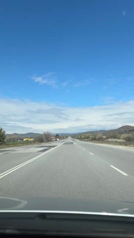 The video shows a first-person perspective of driving on a highway. The road is straight with noticeable repairs and is flanked by dry vegetation and low mountains in the distance. The sky is blue with scattered clouds. Other vehicles are visible on the road ahead. The dashboard of the car from which the video is recorded is visible at the bottom of the frame.