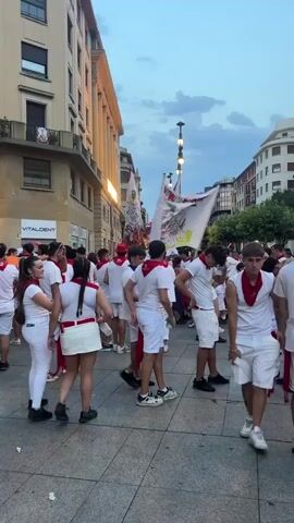 A crowd of people, mostly young adults, are gathered in a street. They are all wearing white clothing, with red sashes or neckerchiefs. Many are holding banners. The buildings on either side of the street are tall and have balconies. The sky is overcast and the light suggests it is late afternoon or early evening. The people appear to be participating in some kind of festival or parade.
