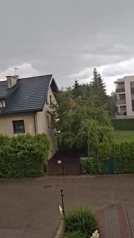 The video shows a residential area during a rain shower. A two-story house with a dark roof is on the left, partially obscured by a dense green hedge. A gate with a sign is visible in front of the house. Behind the house and hedge, a cluster of trees and evergreen bushes create a lush green backdrop. Further to the right, two apartment buildings are visible. The building in the foreground on the right is a multi-story structure with balconies and light-colored walls, accented with a vertical orange panel. The building behind it is similar in design. The sky is overcast with dark, heavy clouds, indicating stormy weather. Raindrops are visible falling throughout the scene, especially noticeable on the paved area in the foreground and on the foliage.