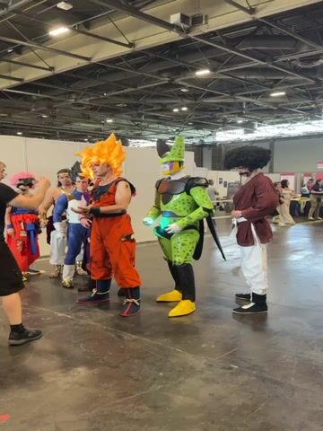 A group of people in cosplay are posing for a photo in what appears to be an indoor convention center. The person in the foreground on the right is dressed as Cell from Dragon Ball Z, wearing a green and black suit with yellow boots and gloves, and holding a glowing green orb. To his left, a person dressed as Goku is wearing his signature orange gi with spiky blonde hair. Behind Goku, a person dressed as Vegeta is wearing a blue and white Saiyan armor. Further back, a person in a red and blue outfit with pink hair is being photographed by someone holding a camera. Another person in the background is dressed in a brown robe with a mustache and afro hairstyle. The environment is a large, open space with a concrete floor and a high ceiling with exposed beams and lighting fixtures.