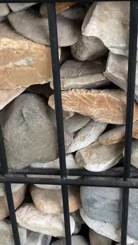The video shows a close-up of a gabion wall, a structure made of a black wire mesh cage filled with various sizes and colors of stones. The stones are predominantly smooth and rounded, with colors ranging from white and grey to tan and brown. The wire mesh has a grid pattern. The camera focuses on the stones and the mesh, and there is no visible movement or interaction within the scene. The background is out of focus.