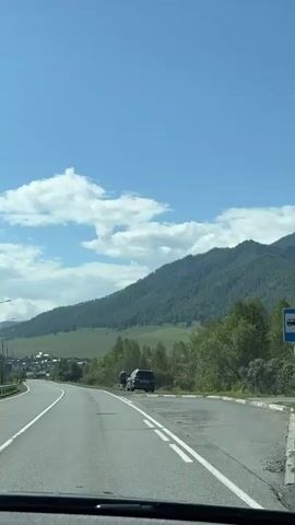 The video shows a car driving on a paved road with a white line marking the lane. To the right of the road, there are some trees and a green mountainous landscape. The sky is blue with scattered white clouds. In the middle of the video, a black SUV is parked on the shoulder of the road. Two people are standing next to the SUV, facing away from the camera. The person on the left appears to be wearing a light-colored shirt and dark shorts, while the person on the right is wearing a dark shirt and shorts. The car in the video continues to drive past the SUV and the two individuals.