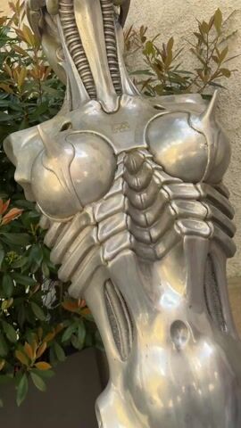 A person touches the breast of a metallic sculpture resembling a female figure. The sculpture has a rib-cage like structure and horn-like projections on the breasts. It has a slender neck with vertical grooves and a head with intricate designs. The sculpture stands beside a green bush against a building. Another person can be seen standing beside the sculpture in the video.
