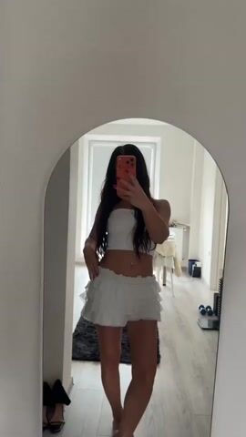A young woman with long dark hair stands in front of a mirror, taking a selfie. She is wearing a white tube top and a short white ruffled skirt. Her arms are crossed and she is looking at the camera. The background is a room with a wooden floor, a rug, and some furniture. There is a radiator on the wall and a window in the background.