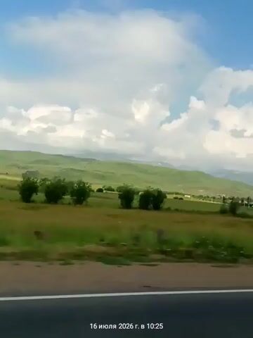The video shows a landscape shot from a moving vehicle, likely a car, as indicated by the blurred road in the foreground and the date and time stamp at the bottom of the screen, which reads "16 июля 2026 г. в 10:25". The environment is rural, with rolling green hills in the background under a bright blue sky dotted with fluffy white clouds. In the middle ground, there are fields of grass and scattered trees, some of which appear to be wind-swept. A red vehicle is briefly visible passing by on the left side of the frame, creating a motion blur. The overall impression is a scenic drive through a countryside.