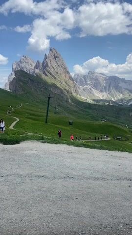 The video shows a mountain landscape with a gravel path in the foreground. A green slope dotted with trees and scattered people stretches towards a jagged, rocky mountain peak under a blue sky with white clouds. A ski lift pole is visible on the slope. In the middle ground, a cluster of buildings, possibly a restaurant or lodge, sits on another green slope. People are seen walking along a path leading to and from the buildings. The overall environment is mountainous and appears to be a scenic area, possibly a hiking or tourist destination during daylight hours.
