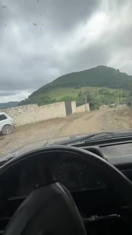 The video begins by looking out from inside a vehicle, focusing on the dashboard and steering wheel. The camera is pointed forward through the windshield, capturing a dirt road winding uphill towards a lush green mountain under a cloudy sky. To the left of the road is a low white brick wall topped with a dark gate. The vehicle appears to be moving slowly, as indicated by the slight blur of the surrounding scenery and the movement of the dashboard needles. The overall impression is a rural, mountainous landscape.