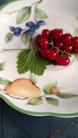 This media file features a close-up shot of a decorative plate with a scalloped green and white rim. The plate is adorned with painted depictions of fruits and leaves, including blueberries, red currants, and what appears to be a peach or apricot. Actual red currants are placed on the plate, positioned over a painted green leaf. The background is a dark, textured surface, possibly a table or cloth. The lighting is soft and diffused, suggesting an indoor setting, possibly in the afternoon or evening. There is no visible text in the media file. The overall impression is one of a still life arrangement, highlighting the artistry of the plate and the fresh berries. The scene evokes a sense of rustic charm and perhaps a culinary theme. The provided location context of Vereya, Russia, adds a geographical anchor to the visual, though it is not directly discernible from the image itself.