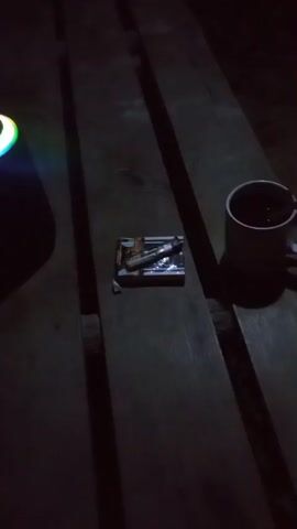 A dark scene of a wooden surface. A cylindrical object with a colorful light ring is visible on the left. In the center, a small, open cigarette pack with a lighter and a cigarette are present. To the right, a dark mug is visible. The lighting is dim, suggesting nighttime or a poorly lit indoor environment.