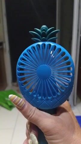 A close-up shot shows a blue, pineapple-shaped, handheld fan being held by a person. The fan has a cage with spokes and a blue pineapple top. The person's hand is visible, with long, decorated fingernails featuring yellow and white stripes. The background is blurred, showing what appears to be a room with some light sources and furniture. The fan appears to be on or in motion, as indicated by slight motion blur.