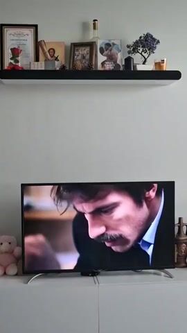 The video shows a television displaying an intimate scene, against the backdrop of a neatly decorated living room wall. On the screen, a man with a dark mustache is initially shown with a pensive expression. The scene then shifts to depict a man holding a woman's face in his hands. The camera angle remains constant, capturing the television and the decorative items on a shelf above it, including framed pictures, a bottle, and a small plant.