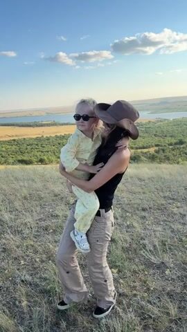 A woman in a black tank top, khaki pants, sneakers, and a brown cowboy hat is holding a young girl in her arms on a grassy hill. The girl is wearing a yellow patterned pajama set and sunglasses. The woman is smiling and has her right arm raised in the air. The background shows a body of water and trees under a blue sky with scattered clouds.