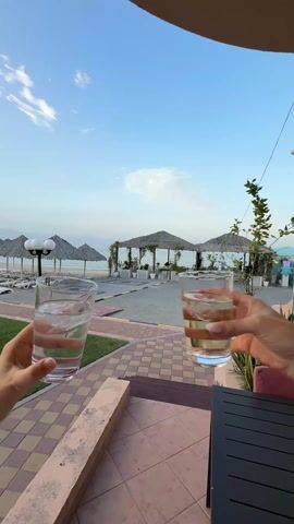 Two hands, one smaller than the other, hold up glasses of water with ice in front of a beach scene. The hands perform a toast with the glasses, one glass having a small splash of orange juice in it. The beach has several tiki-style umbrellas over beach chairs and is edged by a green lawn. The sky is clear blue with light clouds. The person recording is sitting at a black table on a tiled patio.