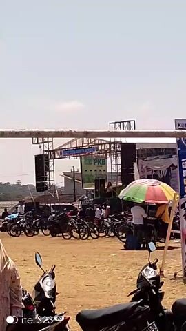 The video shows an outdoor event with a stage set up. On the stage, there is a banner with the letters "PKB" and images of five men. Speakers and sound equipment are visible. In front of the stage, there is a gathering of people and many motorcycles and bicycles parked. Some people are standing and watching the stage, while others are sitting or walking around. The ground is dirt. The sky is clear and bright. There is a colorful umbrella to the right of the frame. In the foreground, two motorcycles are parked, and to the left, a person wearing a headscarf is visible. A child in a white shirt and cap is also in the foreground.