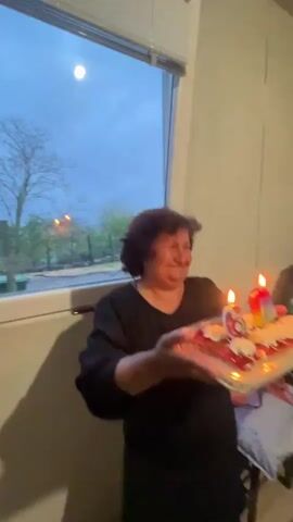 In a heartwarming indoor scene in Kosten, Bulgaria, an older woman with dark, short hair holds a rectangular birthday cake adorned with lit candles. She wears a black top and smiles broadly, her face radiating joy. The cake features a red jelly or fruit topping with white cream decorations, and prominently displays a number "6" candle alongside another colorful candle, both burning brightly.

Behind the woman is a large window, through which the dim light of dusk or early night is visible, with silhouettes of bare trees and distant lights. The room itself has light-colored walls. In the background, a smartphone rests on a table, next to what appears to be a laptop or computer monitor. The sounds of clapping are audible, suggesting a celebration, likely a birthday party.
