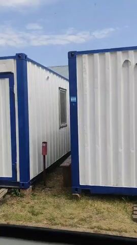 A camera pans across a row of modular shipping container-style buildings. The first building has a white exterior with blue trim and a white door with a green sign. Next to it is another similar building, followed by a longer building with a window. The camera continues to pan, showing more containers, some with air conditioning units mounted on the exterior. The ground is covered in dirt and grass, and the sky is visible with some clouds. The scene appears to be an outdoor storage or temporary housing area.
