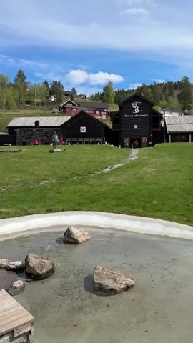 The video shows a series of outdoor scenes in a scenic location. 

The first scene focuses on a small, circular pond with several large rocks within it.  Behind the pond is a grassy area leading up to a series of dark wooden buildings, one of which is labelled "Norsk Skieventyr" on its facade. A statue of a person is standing in the grass near the buildings. The sky is blue with scattered clouds.

The camera pans to the left, revealing a parking area with multiple vehicles. In the background, there are houses, trees and rolling hills.

The next shot shows a miniature cabin replica on a wooden platform, with a lake visible in the background.

The final shot shows a small, rocky island in the middle of the lake. A metal sculpture resembling a bowl rests atop the pile of rocks. The surrounding landscape consists of mountains, trees and small buildings. The water is calm and reflects the sky and the island.