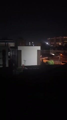 This is a nighttime video of an urban or suburban landscape. Buildings are visible, some illuminated by artificial light, others in shadow. A dark foreground suggests the viewpoint is from a window or balcony. The sky is dark, indicating it is late at night or early morning. There are no people or discernable actions visible in the video.