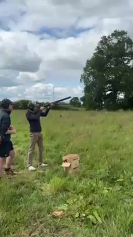 Here is a description of the video:

The video begins with a man standing in a grassy field, holding a shotgun. He's wearing a dark jacket, tan pants, and what appear to be hearing protectors. Next to him is a cardboard box. The man raises the shotgun, aims, and fires. The video then shows a clay pigeon target being launched into the air from a mechanical trap. The background includes green trees and a cloudy sky. The setting is outdoors in a rural area.
