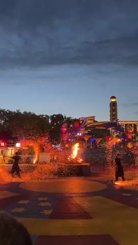 Two performers in dark clothing are engaged in a fire show in an outdoor plaza at dusk. They are using fire-twirling devices that create arcs of bright orange sparks. In the center of the plaza, a central fire feature is also producing sparks. The plaza is decorated with colorful flags and structures, and trees are visible in the background. A building with a prominent tower is also in the distance. The sky is a deep blue with some clouds. People are observing the performance.