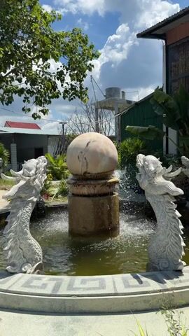 The video shows a garden with a fountain in the center. The fountain has a large sphere on top and is surrounded by dragon statues. There are also various plants and trees in the garden, including palm trees and banana trees. The video is shot from a fixed perspective and pans slowly to the right. The weather appears to be sunny with some clouds in the sky.