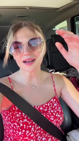 The video shows a blonde woman in a red floral dress and sunglasses sitting in the driver's seat of a car. Her mouth is open as if she is singing or talking. Her left hand is raised near her head, and her right hand is blurred in motion, possibly waving. The interior of the car is visible, including a seatbelt, dashboard, and the headrest of the passenger seat. The background appears to be outdoors, with sunlight visible through the car windows.