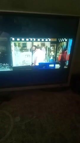 The video shows a television screen. The screen displays several scenes, including two men walking past a shop window, a woman sitting at an outdoor cafe and looking at her phone, and a man standing in front of a body of water. The room is dark. The television sits on a patterned surface.