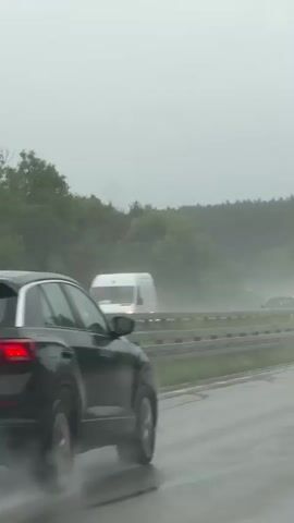 The video shows a black Volkswagen T-Roc driving on a highway in heavy rain. The car is kicking up a lot of spray from the wet road, obscuring the rear of the vehicle. Other cars are visible in the distance, also driving through the spray. The surrounding environment appears to be a forest, with trees lining the highway. The sky is overcast and gray, indicating the heavy rainfall. The video is shot from a moving vehicle, likely following the T-Roc.