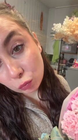 A woman stands in a flower shop and holds a bouquet of pink and green carnations wrapped in pink and white paper. Her hair is long and brown, and she wears a gray zip-up sweater and a black apron. The background includes other floral arrangements, a ladder, and shop fixtures. The lighting is bright, and the video appears to be shot indoors. The woman makes a kissing face towards the camera.