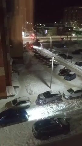 The video shows a nighttime scene of a snow-covered parking lot and street in a city. The view is from an elevated position, likely a window in a building. There are several cars parked and covered in snow. Streetlights illuminate the area. In the distance, there are apartment buildings. A person walks on the sidewalk in the background. Several cars move on the street. The video is a bit shaky, with some blurring.