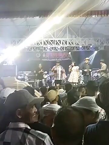 A concert is taking place outdoors under a large tent. A band is performing on a raised stage with bright lights. The band consists of at least six musicians, including a female singer in the center wearing a light-colored patterned dress. She is holding a microphone and appears to be singing. The other band members are playing instruments such as guitars and drums. Behind the band, a large banner reads "TOM MORENSA". A crowd of people is gathered in front of the stage, many of them holding up their hands or phones. The audience is densely packed and appears to be enjoying the performance. The lighting is dim, with spotlights illuminating the stage. The overall atmosphere seems lively and energetic.