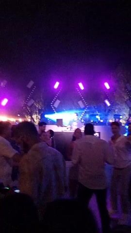 A group of people are dancing at a night club. They are in front of a stage with a DJ. The stage is lit with blue and pink lights. The people are dressed in casual clothing. Some of them are holding phones and recording the performance.