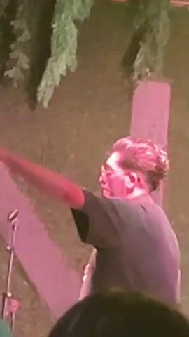The video shows a man with glasses and dark curly hair, wearing a dark t-shirt, standing on a stage at night. He is holding a microphone and appears to be singing or speaking. The stage is lit with pink and green lights, and there are trees in the background. Another man, wearing sunglasses and a dark shirt, is also visible on stage, holding a microphone. The audience is not clearly visible, but there are people in the foreground, some of whom appear to be holding up their hands. The overall atmosphere seems to be a live performance or concert.