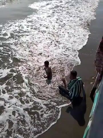A child is playing in shallow water at the edge of a beach. Waves are washing onto the shore around the child. A person wearing a striped shirt is sitting on the sand near the child, facing away from the camera. Another person, partially visible on the right, is standing at the edge of the frame. The sun is shining, creating bright reflections on the water and wet sand.