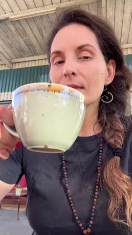 The video shows a close-up of a woman with long, dark hair braided to her side. She is wearing a black t-shirt and a beaded necklace. She is holding a light green mug with a beige rim and drinking from it. Her right hand is holding the mug. Her left hand is visible on the bottom left of the screen. She lowers the mug and then sticks her tongue out at the camera. The background appears to be an outdoor covered patio with a corrugated metal wall.