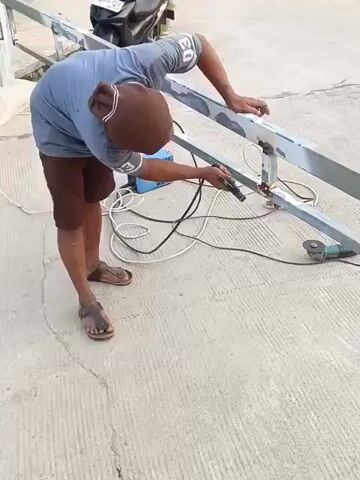 A person is welding a metal structure outdoors. They are wearing a blue t-shirt, brown shorts, and sandals. The person is bent over, holding a welding torch to the metal. Sparks are flying from the welding point. A blue welding machine is on the ground behind the person, connected by cables. A metal grinder is also visible on the ground near the welding area. The welding is taking place on a concrete surface, possibly a walkway or bridge, with a metal railing or fence being worked on. A motorcycle is parked in the background. The scene appears to be during daylight.