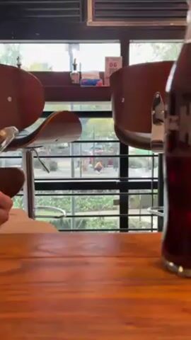 The video shows a man doing squats outside. The scene is viewed through the window of what appears to be a cafe or restaurant. There is a table and chairs in the foreground, and a bottle of dark liquid. Outside, the man is shirtless and wearing dark pants and white sneakers. He is performing squats next to a metal structure. A red car is parked behind him. The view is partially obscured by the window frame.
