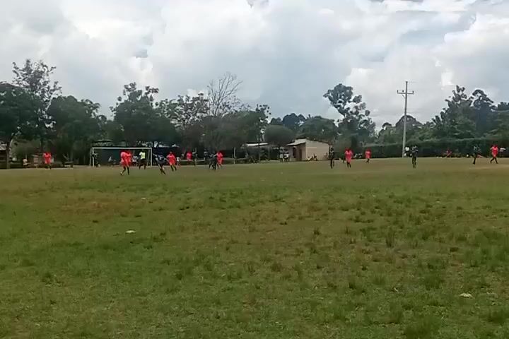 A soccer match is in progress on a grass field. There are multiple players on the field wearing orange and black uniforms. The game is in motion with players actively running and pursuing the ball. The field is surrounded by trees and some simple structures can be seen in the background. The sky is overcast with some cloudy patches.  The overall atmosphere suggests a casual, possibly amateur, sports event taking place in a rural or suburban setting.
