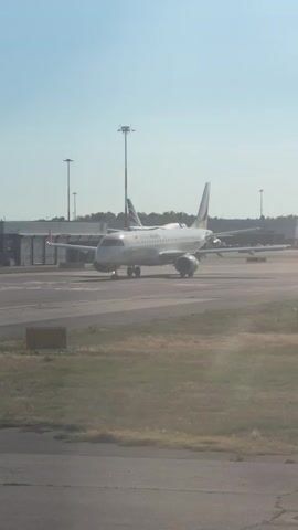 An airplane is on a tarmac at an airport. The plane is white with yellow and red markings on its tail and fuselage. It has two engines under its wings and is facing the left of the frame. In the background, there are airport buildings, tall light poles, and other parked airplanes. The sky is clear and blue, and the ground is covered with pavement and some grass. The camera is positioned at a low angle, looking up at the plane. The video is brief, showing the plane stationary on the tarmac.