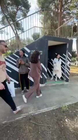 The video captures a group of people entering a small, bunker-like structure with the Hebrew word "Miklat" written on it, meaning shelter. A man wearing a beige sweatshirt and black pants with a bag over his shoulder leads the group, followed by a woman in a brown outfit. Two women in white outfits exit the bunker just before the man enters. Another person with long, dark hair is standing outside, using a cell phone, wearing a grey jacket with yellow stripes on the pants. The structure is painted black with white stripes and has stairs leading to the roof. The area appears to be outdoors, with trees and greenery visible in the background.