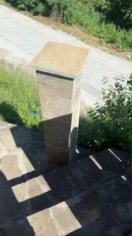The video pans from a stone pillar and steps to a lush green forest on a sunny day. The pillar is light brown and square, with a flat top. The steps are dark gray and appear to be made of stone. A winding road is visible in the background, lined with trees and foliage. The sky is a clear blue.
