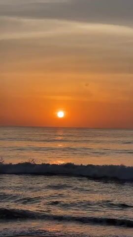 The video shows a serene ocean sunset. The sun is a bright, small orb positioned just above the horizon, casting a warm orange glow across the sky and reflecting on the water. Gentle waves roll towards the shore, creating a soothing rhythm. The sky transitions from a deep orange near the sun to lighter shades of orange and gray further up. The overall atmosphere is peaceful and tranquil.